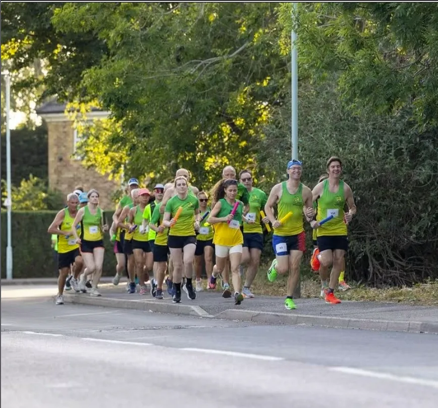 A big group of club runners in green and gold vests running along a tree-lined road, some carrying relay batons