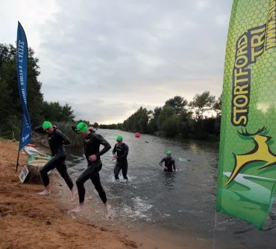 Swimmers in wetsuits entering a lake at a local triathlon, next to a Stortford Tri flag